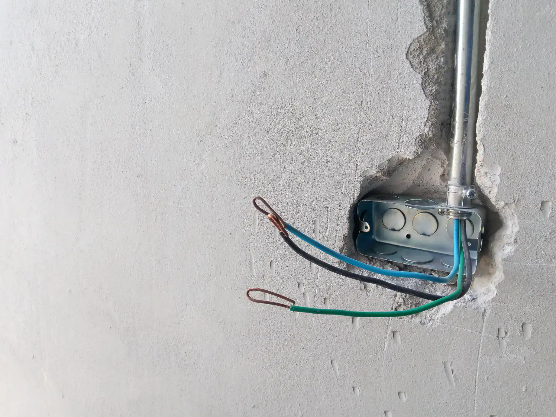 An electrical junction box mounted in a wall with three exposed wires&mdash;black, green, and blue&mdash;protruding from it. The wires have looped ends, and the unfinished wall suggests ongoing work by electricians in Rhode Island.