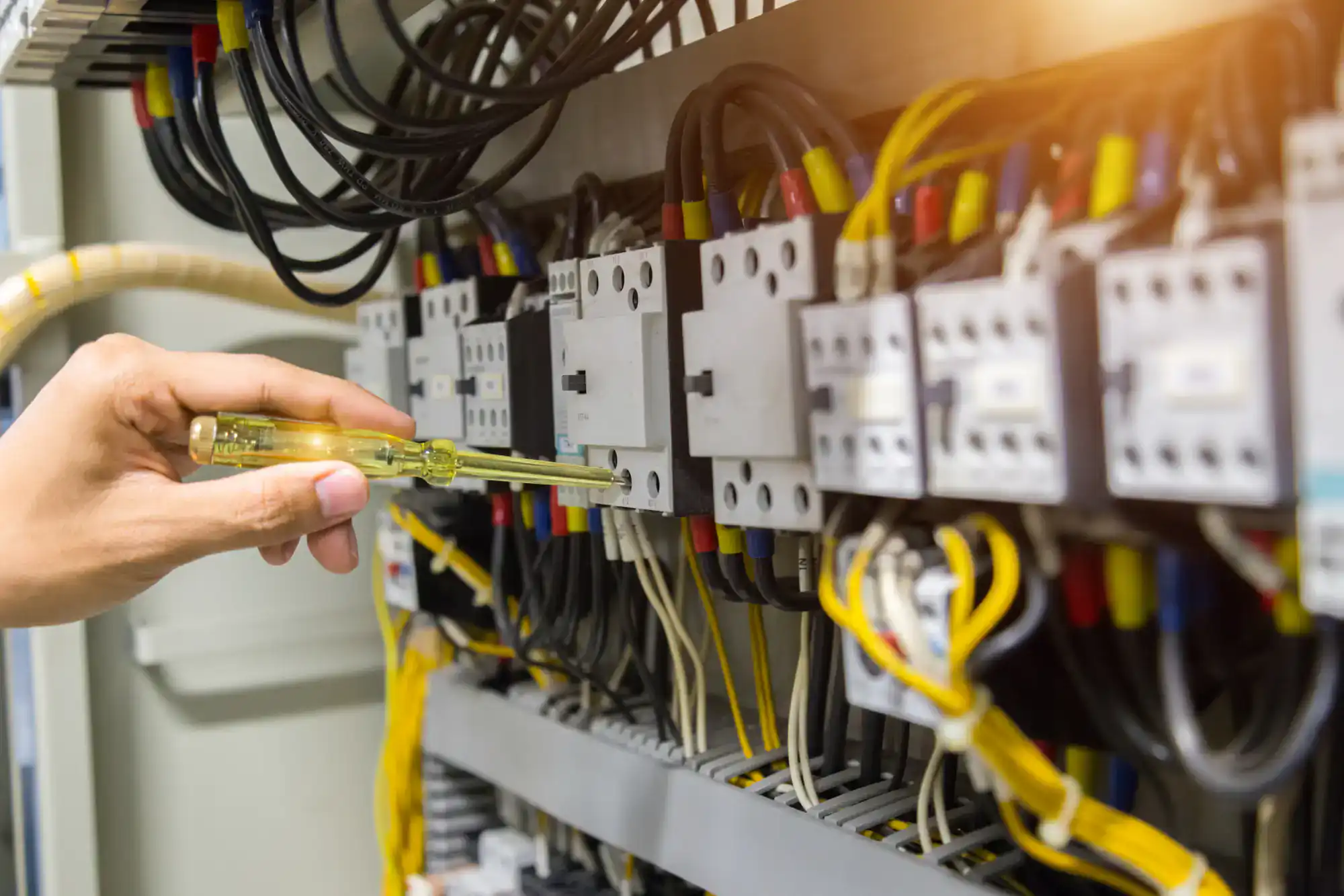 A hand holding a screwdriver tests or repairs an electrical control panel filled with wires, switches, and circuit components—just the kind of work skilled electricians in Rhode Island handle daily.