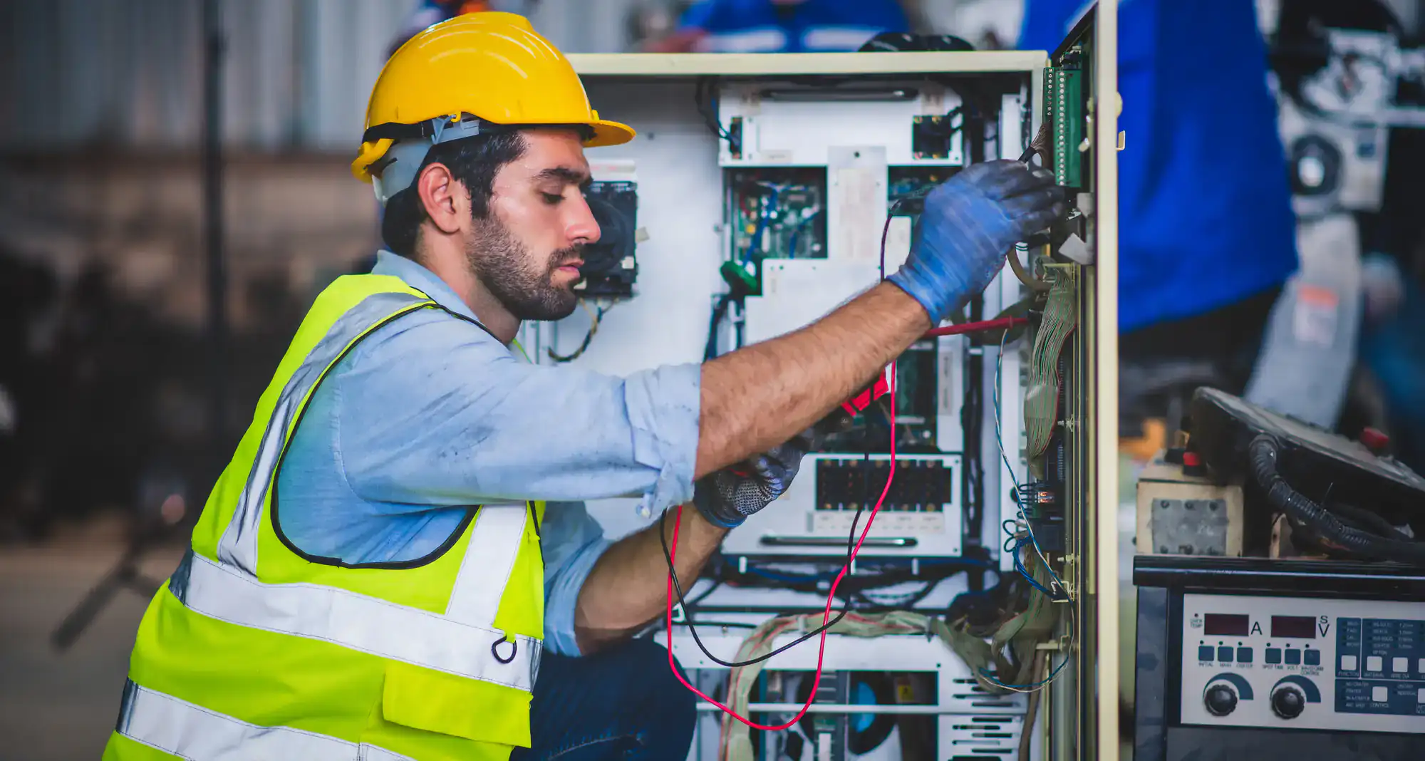 A man wearing a yellow safety vest, helmet, and gloves is repairing or inspecting electrical components inside an industrial control panel. He is holding test probes and working attentively, typical of skilled electricians Rhode Island RI.
