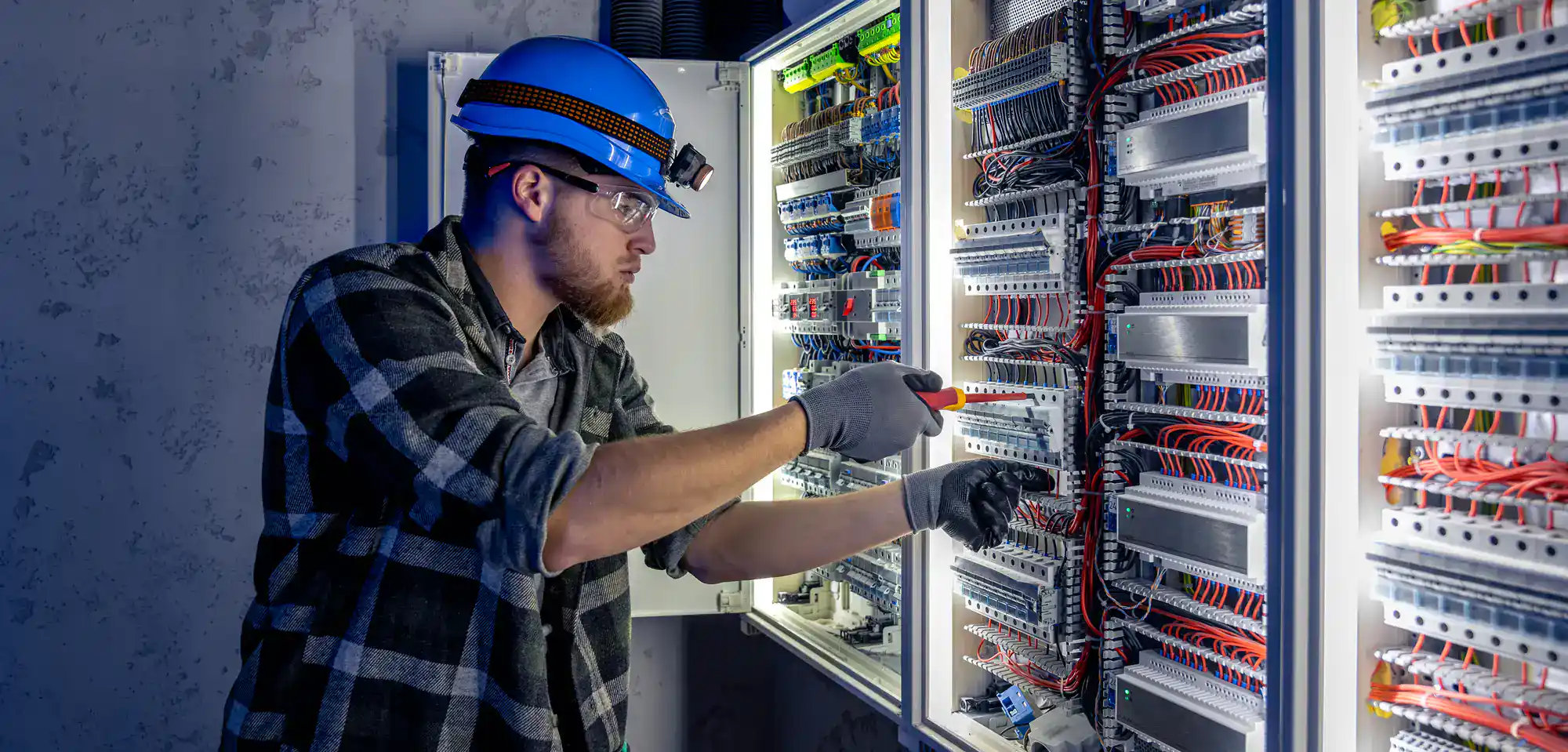 An electrician in Rhode Island, wearing safety gear, works with wires and components inside an open electrical control panel, using a screwdriver and carefully inspecting the connections.