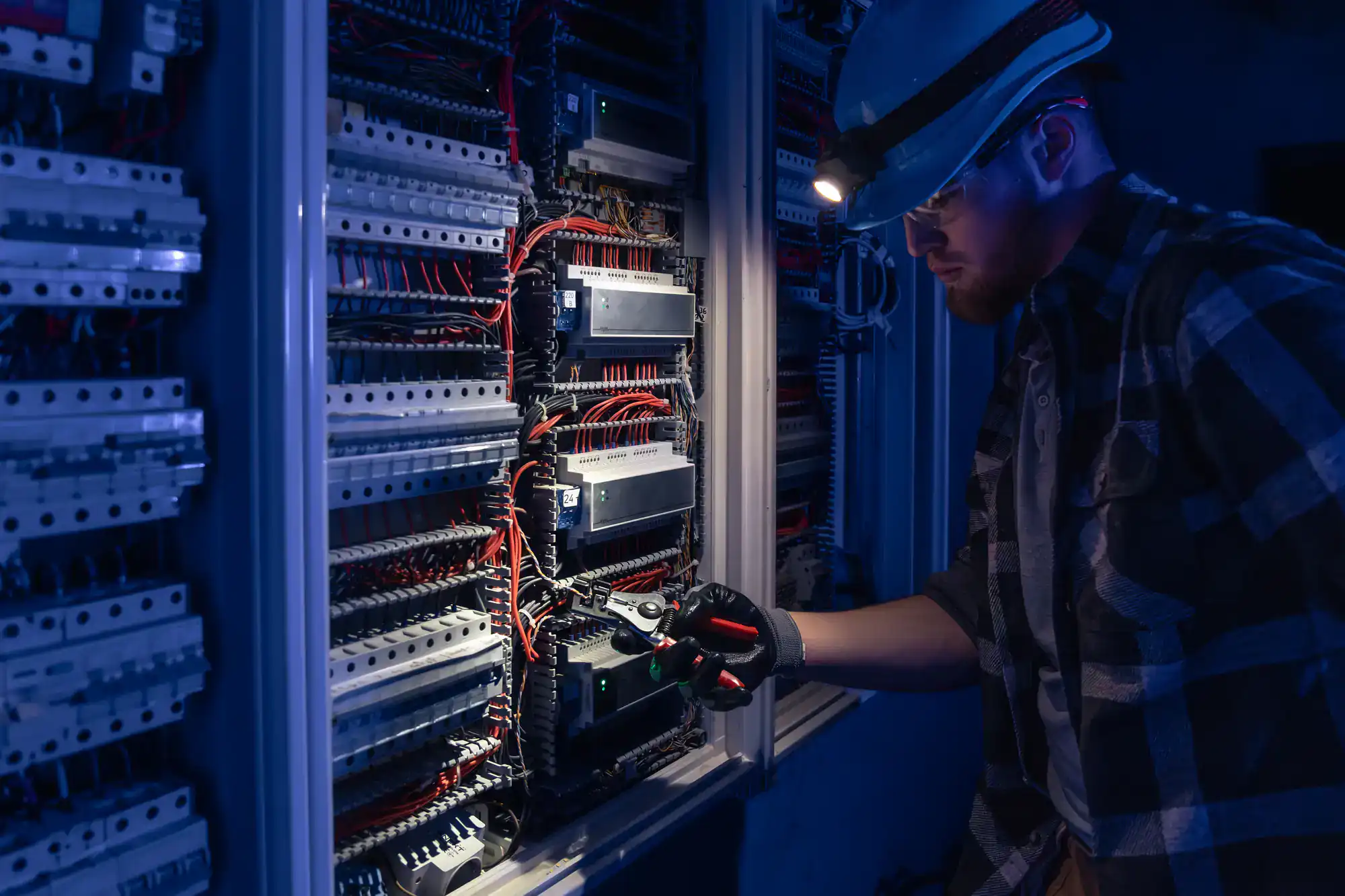 An electrician from electricians Rhode Island, wearing a helmet and headlamp, inspects and works on a large electrical panel with numerous wires and circuit breakers in a dimly lit setting.