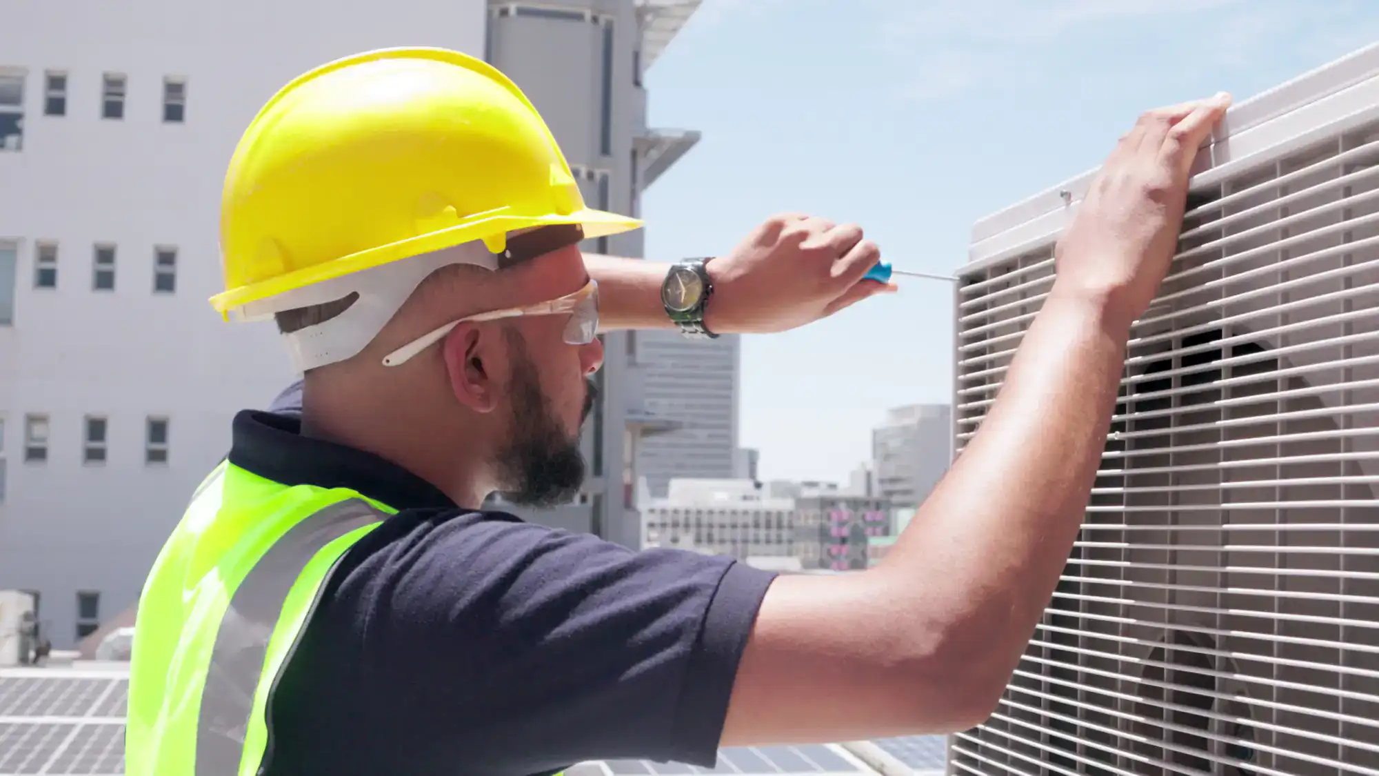 A worker wearing a yellow hard hat, safety glasses, and a neon vest repairs an outdoor air conditioning unit on a rooftop in Rhode Island, possibly one of the local electricians, with buildings visible in the background.